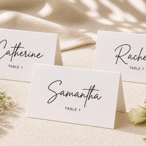 Könnte beinhalten: Drei weiße Tischkarten mit eleganten schwarzen Kalligraphie-Namen: "Catherine", "Rachel" und "Samantha". Jede Karte zeigt auch eine Tischnummer. Die Karten sind auf einer strukturierten Oberfläche angeordnet, mit weißen Blumen im Vordergrund.