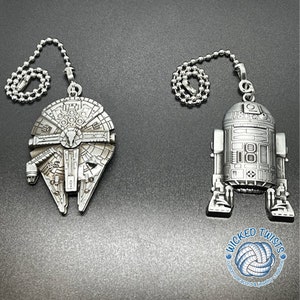 May include: Two silver metal fan pull chains, one shaped like the Millennium Falcon spaceship and the other shaped like the R2-D2 robot from Star Wars. The chains are attached to a silver metal ring with the text "Wicked Twists Custom Paracord & Jewelry Creations".
