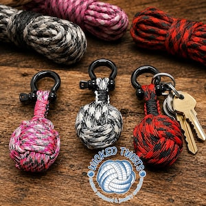 May include: Three paracord keychains in pink, black and white, and red and black patterns. Each keychain has a black metal shackle and a woven knot design. The keychains are displayed with spools of matching paracord.