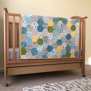May include: A wooden crib with a yellow, blue, and gray patterned quilt. The crib has a drawer underneath.