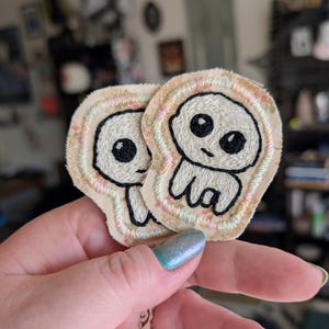 May include: Two embroidered patches featuring a cartoon-like character with large black eyes and a simple outline. The patches are beige with a floral border and are held in a hand. The character's design is in black thread.