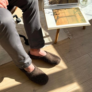 May include: A pair of brown wool slippers with a fuzzy texture. The slippers are worn by a person sitting on a chair.