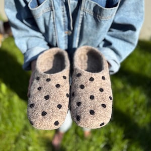 Könnte beinhalten: Ein Paar beige Filzschuhe mit schwarzen Tupfen. Die Hausschuhe sind weich und scheinen handgefertigt zu sein. Das Schuhwerk ist für den Innenbereich konzipiert und bietet Komfort und Wärme.
