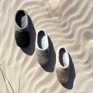 May include: Three pairs of wool slippers in different shades of gray. The slippers are on a sandy surface.