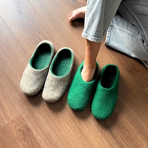 May include: Two pairs of felted wool slippers. One pair is light gray with green interior, the other is bright green. The slippers are on a wooden floor, with a person's foot entering one of the green slippers.