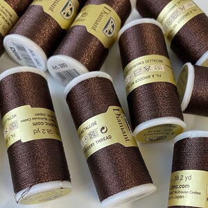 Op de afbeelding: Een close-up van meerdere spoelen met bruine metallic borduurgaren. De spoelen zijn geëtiketteerd met "Diamant" en hebben de tekst "Metallic Embroidery Thread" en "Fil à Broder Métallique" erop gedrukt. De spoelen zijn gemaakt in Japan en zijn 35 meter lang.