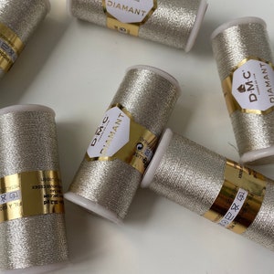Op de afbeelding: Zes spoelen DMC Diamant metallic borduurgaren in zilverkleur. De spoelen zijn in een cirkel op een wit oppervlak gerangschikt. De spoelen hebben een gouden label met het DMC-logo en de tekst "Diamant" in zwart.