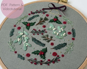 May include: An embroidered hoop with a Christmas wreath design. The design features green and red embroidered leaves and berries, including holly and mistletoe, on grey fabric. The text "PDF Pattern & Videotutorial" is visible.