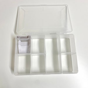 Può includere: Una scatola organizer in plastica trasparente e rettangolare con coperchio incernierato, divisa in più scomparti. La scatola contiene piccole carte bianche con un disegno circolare. Il coperchio è aperto.