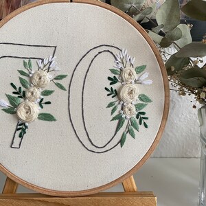 Op de afbeelding: Geborduurd kunstwerk op een rond canvas, met het nummer "70" met bloemversieringen. De "7" en "0" zijn omlijnd met zwarte draad en versierd met cr&egrave;mekleurige rozen en groene bladeren. Het canvas is geplaatst in een houten hoepel.