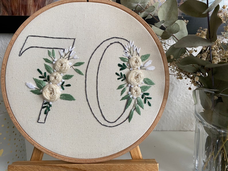 Op de afbeelding: Geborduurd kunstwerk op een rond canvas, met het nummer "70" met bloemversieringen. De "7" en "0" zijn omlijnd met zwarte draad en versierd met cr&egrave;mekleurige rozen en groene bladeren. Het canvas is geplaatst in een houten hoepel.