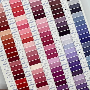 Op de afbeelding: Een close-up van een kleurenkaart met rijen garenrollen. De rollen zijn gerangschikt in rijen per kleur, beginnend met rood en overgaand naar paars. Elke rol heeft een gedrukt nummer.