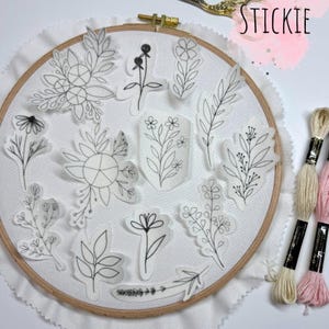 Könnte beinhalten: Eine Sammlung von Aufklebern mit floralen Motiven, darunter Blätter und Blumen, angeordnet auf weißem Stoff in einem Stickrahmen. Das Wort "STICKIE" ist in Pink geschrieben. Stickgarn ist sichtbar.