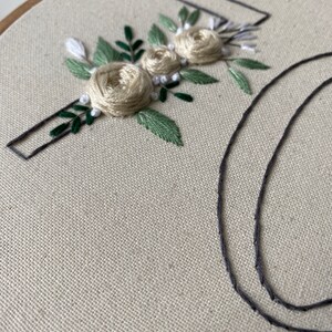 Op de afbeelding: Geborduurd bloemmotief op een beige stof gespannen in een houten hoepel. De borduursels tonen cr&egrave;mekleurige rozen, groene bladeren en witte accenten. Een donkergrijze letter "J" is boven de bloemen geborduurd, met een gedeeltelijke "O".