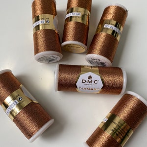 Op de afbeelding: Zes spoelen goudkleurig metallic garen. De spoelen zijn gelabeld met "DMC Diamant" en "Fil a Broder Metallic."