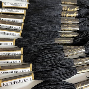 May include: Close-up of several skeins of black embroidery floss. Each skein is wrapped with a paper label that includes text and a barcode. The floss is made of 100% cotton. The image shows a collection of craft supplies.
