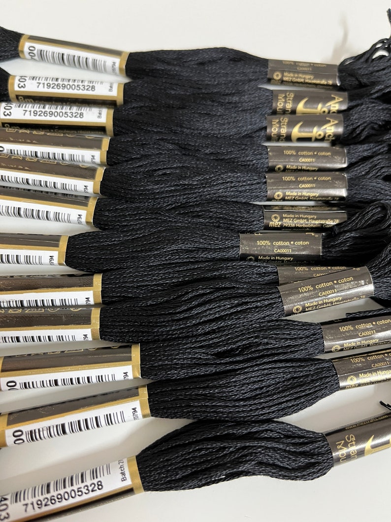 May include: Close-up of several skeins of black embroidery floss. Each skein is wrapped with a paper label that includes text and a barcode. The floss is made of 100% cotton. The image shows a collection of craft supplies.
