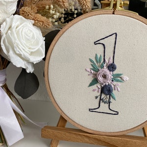 Op de afbeelding: Geborduurd nummer &eacute;&eacute;n in een houten ring, versierd met bloemaccenten in tinten lavendel, blauw en groen. De borduurring wordt weergegeven op een klein houten ezel, met een boeket witte rozen en droogbloemen op de achtergrond.
