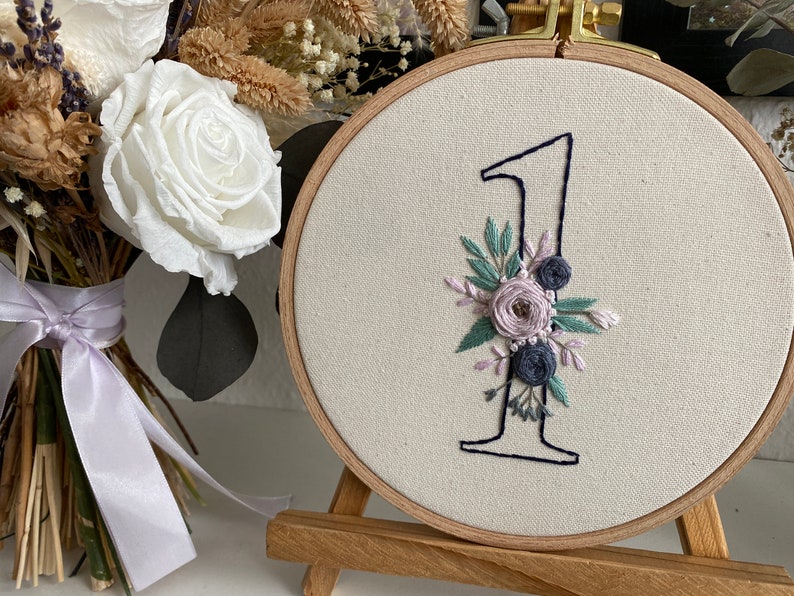 Op de afbeelding: Geborduurd nummer &eacute;&eacute;n in een houten ring, versierd met bloemaccenten in tinten lavendel, blauw en groen. De borduurring wordt weergegeven op een klein houten ezel, met een boeket witte rozen en droogbloemen op de achtergrond.