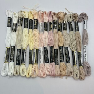 Può includere: Una collezione di fili da ricamo Anchor in varie tonalità pastello, tra cui bianco, crema, giallo, rosa e beige. Ogni matassa, del marchio Anchor, misura 8 metri di lunghezza. I fili sono disposti per mostrare la varietà di colori.