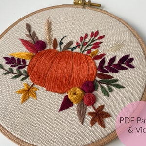 Könnte beinhalten: Stickrahmen-Kunst mit Stickerei, die einen Kürbis mit Herbstblättern und Blumen in Orange-, Rot-, Gelb- und Grüntönen zeigt. Der Rahmen ist aus Holz und hat einen natürlichen Leinenstoff.