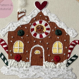 Puede incluir: Casa de jengibre bordada con bastones de caramelo, un corazón rojo y un hombre de jengibre en la ventana. La casa está rodeada de nieve blanca y acebo verde.