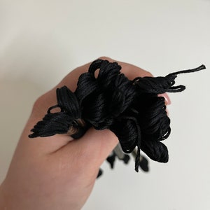 May include: A bundle of black embroidery floss held in a hand. The floss is tightly wound and appears to be made of a smooth, shiny material. The threads are a deep, solid black color.