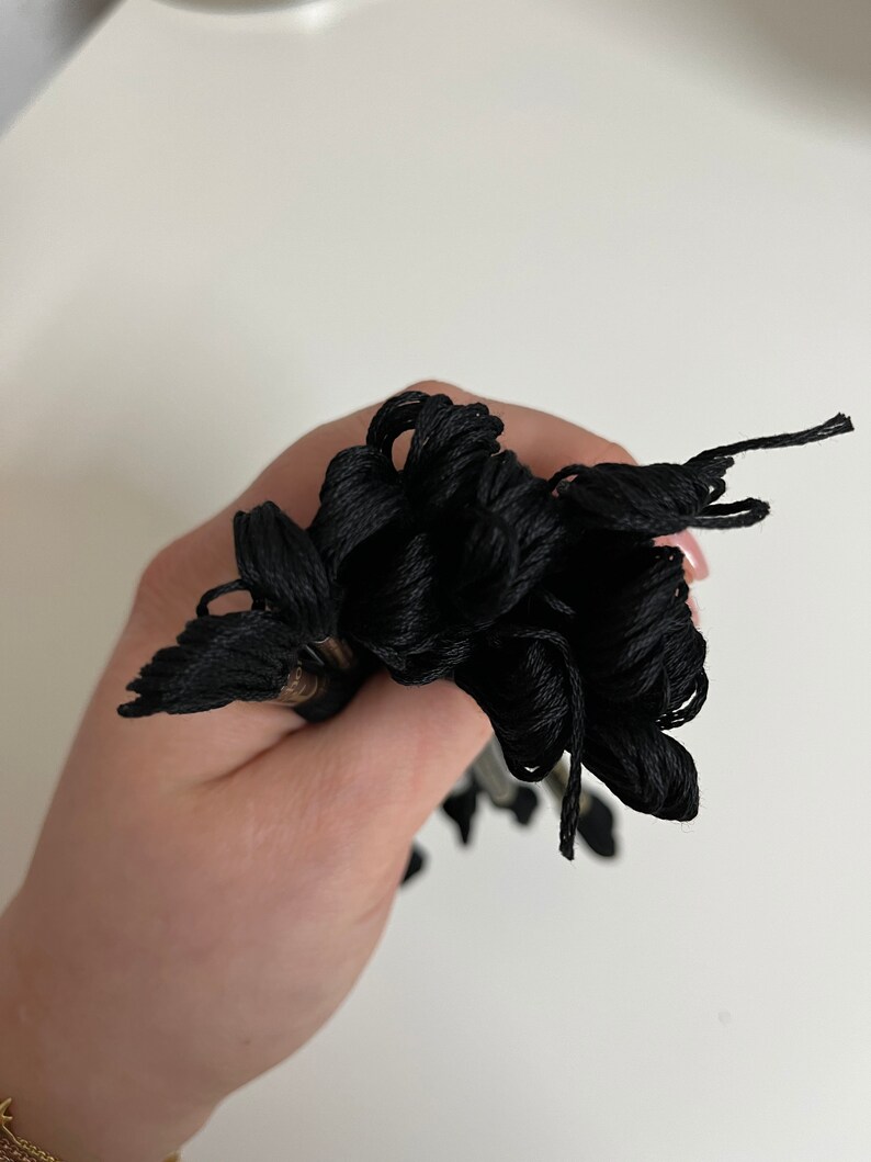 May include: A bundle of black embroidery floss held in a hand. The floss is tightly wound and appears to be made of a smooth, shiny material. The threads are a deep, solid black color.