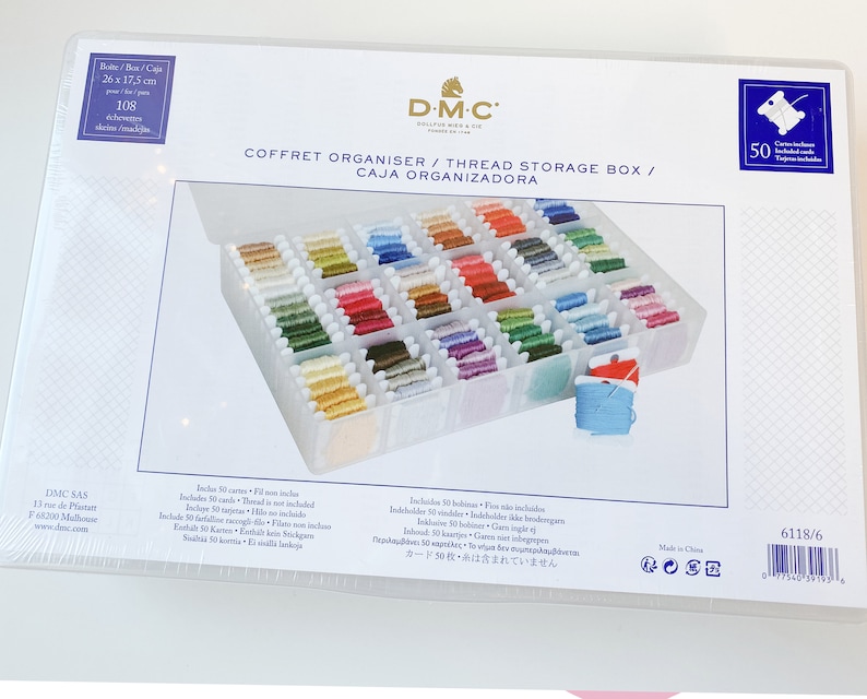 May include: A clear plastic thread storage box with a white lid. The box contains numerous spools of colorful embroidery floss. The box is labeled "COFFRET ORGANISER / THREAD STORAGE BOX / CAJA ORGANIZADORA" and measures 10.2 x 6.9 inches.