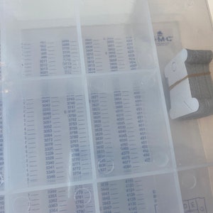 May include: A clear plastic organizer with multiple compartments, each labeled with numbers. A stack of small, gray metal tags is secured with a rubber band in the upper right corner. The organizer is likely for small parts.