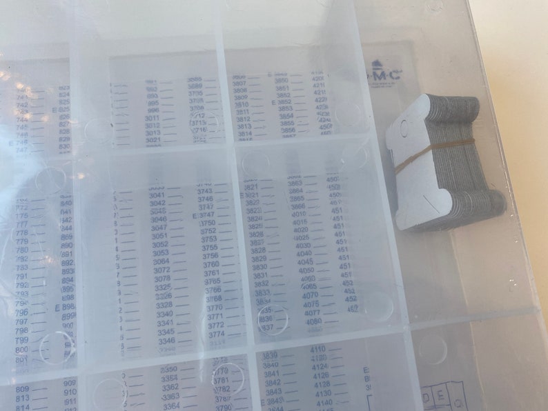 May include: A clear plastic organizer with multiple compartments, each labeled with numbers. A stack of small, gray metal tags is secured with a rubber band in the upper right corner. The organizer is likely for small parts.