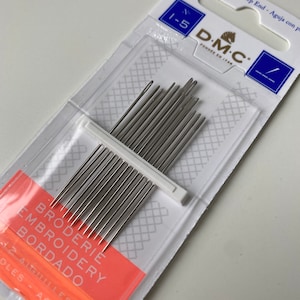 Op de afbeelding: Een verpakking met 12 DMC borduurnaalden, maat 1-5, met een witte kunststof houder. De verpakking is wit met blauwe en rode accenten. De tekst op de verpakking luidt "Bout pointu - Sharp End - Aguja con pa N 1-5 DMC FONDÉE EN 1746 BRODERIE EMBROIDERY BORDADO 12 AIGUILLES DLES - AGUJAS".