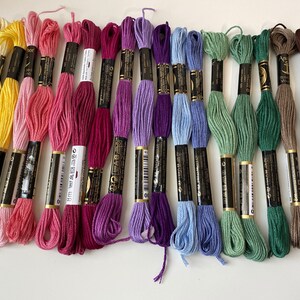 Könnte beinhalten: Sortiment von 18 Stickgarn-Spulen in verschiedenen Farben, darunter Gelb, Rosa, Lila, Blau, Grün, Braun und Grau. Jede Spule hat ein schwarzes Etikett mit goldenen Buchstaben.