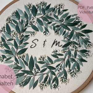 May include: A white embroidery hoop with a green leafy wreath design and the letters "S & M" in the center. The text "PDF Pattern & Videotutorial" is in the upper right corner and the text "Alphabet enthalten" is in the lower left corner.