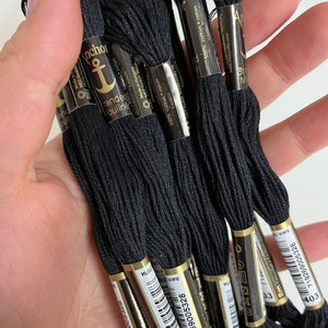 May include: A collection of black embroidery floss skeins, held in a hand. Each skein is labeled with the brand "Anchor" and includes details like "100% cotton" and "8m". The floss is a deep, solid black color, suitable for various needlework projects.