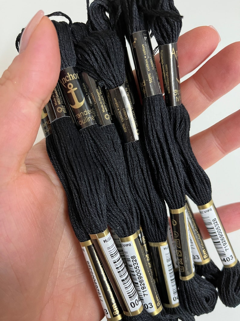 May include: A collection of black embroidery floss skeins, held in a hand. Each skein is labeled with the brand "Anchor" and includes details like "100% cotton" and "8m". The floss is a deep, solid black color, suitable for various needlework projects.