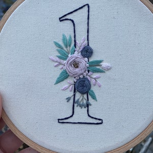 Op de afbeelding: Geborduurde nummer &eacute;&eacute;n in marineblauw, versierd met een bloemstuk in tinten lavendel, saliegroen en grijs. De borduursel is geplaatst in een houten hoepel, waardoor een decoratief stuk ontstaat.