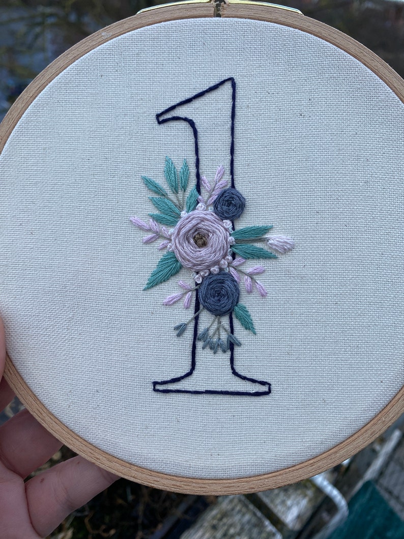 Op de afbeelding: Geborduurde nummer &eacute;&eacute;n in marineblauw, versierd met een bloemstuk in tinten lavendel, saliegroen en grijs. De borduursel is geplaatst in een houten hoepel, waardoor een decoratief stuk ontstaat.