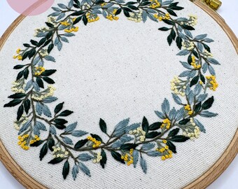 May include: A circular embroidery hoop with a white fabric background. The fabric is embroidered with a wreath of green and blue leaves and yellow flowers.