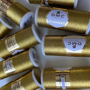 Könnte beinhalten: Nahaufnahme von mehreren Spulen goldfarbenen Metallic-Garn. Die Spulen sind mit "DMC Diamant" beschriftet und zeigen ein Pferdelogo. Das Garn wird in Frankreich hergestellt.