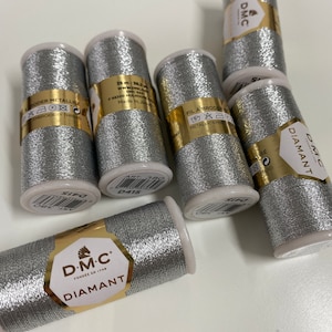 Op de afbeelding: Zes spoelen zilveren metallic borduurgaren met gouden etiketten. De etiketten dragen de tekst "DMC Diamant" en "Fil à Broder Metallic Embroidery Thread".