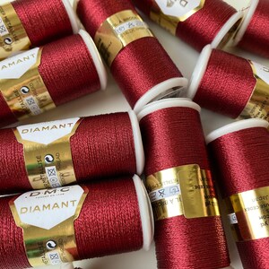Op de afbeelding: Een verzameling bordeauxrode metallic borduurgaren spoelen. Elke spoel heeft een witte basis en een gouden label met de merknaam "DIAMANT". Het garen is strak opgerold en de spoelen liggen verspreid over een wit oppervlak.