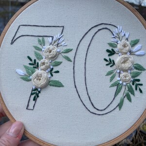 Op de afbeelding: Geborduurde borduurringkunst met het nummer 70. De cijfers zijn omlijnd met donkergrijze draad en versierd met cr&egrave;mekleurige rozen, witte accenten en groene bladeren. De borduursels zijn geplaatst op een neutrale stoffen achtergrond, omlijst door een houten ring.