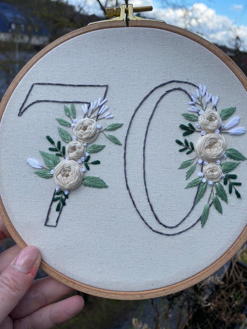 Op de afbeelding: Geborduurde borduurringkunst met het nummer 70. De cijfers zijn omlijnd met donkergrijze draad en versierd met cr&egrave;mekleurige rozen, witte accenten en groene bladeren. De borduursels zijn geplaatst op een neutrale stoffen achtergrond, omlijst door een houten ring.