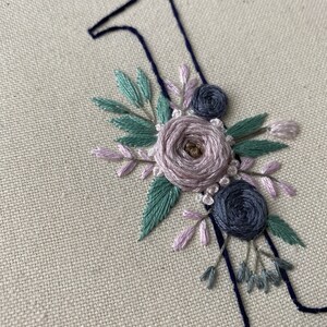 Op de afbeelding: Geborduurde nummer &eacute;&eacute;n met bloemaccenten. Het nummer is donkerblauw en de bloemen zijn lavendel, saliegroen en donkerblauw. De borduursels zijn op een beige stof in een houten ring.