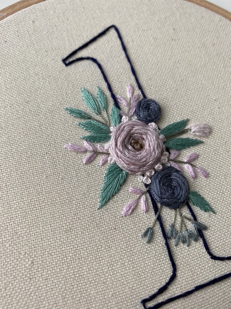 Op de afbeelding: Geborduurde nummer &eacute;&eacute;n met bloemaccenten. Het nummer is donkerblauw en de bloemen zijn lavendel, saliegroen en donkerblauw. De borduursels zijn op een beige stof in een houten ring.
