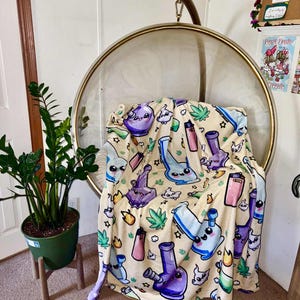 May include: A beige blanket with a repeating pattern of cartoon bongs, lighters, and marijuana leaves in purple, blue, and pink. The blanket is draped over a clear, round hanging chair.