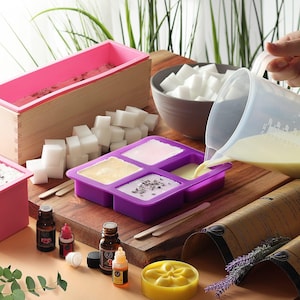May include: A purple silicone mold with four sections, filled with a white liquid. There are small wooden sticks, essential oil bottles, and a wooden box with a pink silicone mold inside. There are also white soap cubes and a bowl of white soap cubes.