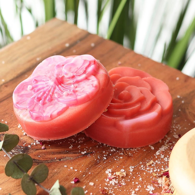 May include: Two pink soap bars with floral designs on a wooden surface. The soap bars are shaped like a flower and a rose.
