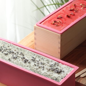 May include: Two pink silicone molds for making soap. One mold is filled with a light green soap with dried lavender buds. The other mold is filled with a pink soap with dried rose petals.